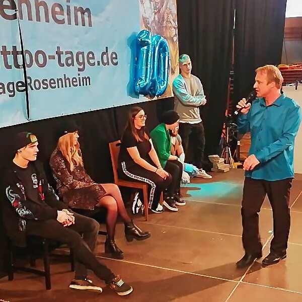 KUBUDO spricht in ein Mikrofon vor mehreren sitzenden und stehenden Personen auf einer Bühne, während ein Banner mit "Tattoo-tage.de" im Hintergrund zu sehen ist.