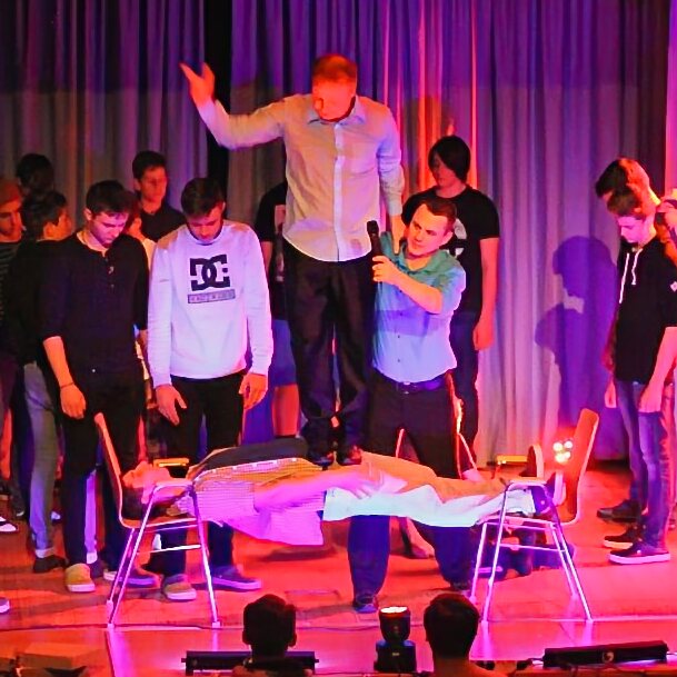 Hypnotisierter junger Mann liegt in kataleptischer Starre zwischen zwei Stühlen, KUBUDO steht auf seinem Körper